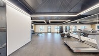 Located at 1002 Niagara St in Welland, Ontario, this modern fitness facility offers a thoughtfully designed exercise room that provides a comfortable and functional environment for a variety of workout routines. The space features a striking contemporary aesthetic, defined by a bold dark ceiling with sleek linear LED lighting strips that create a clean, energizing atmosphere throughout.

The exercise room is finished with durable rubber flooring, ideal for high-impact activities, and benefits from generous natural light through multiple windows and a glass-paneled exterior door that opens to an outdoor balcony area with views of the surrounding neighbourhood. The bright white walls contrast effectively with the dark ceiling, contributing to an open and inviting feel.

Equipment is well-distributed throughout the space and includes cardio machines such as treadmills, a multi-station cable machine, and an assortment of free weights including a full dumbbell rack paired with an adjustable bench. A wall-mounted flat-screen TV adds convenience for entertainment during workouts. Decorative fitness-themed wall graphics add personality and reinforce the athletic character of the room. Access is controlled via a key fob entry system, ensuring a secure environment for users. Overall, this facility delivers a polished and fully functional fitness experience.