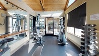 This fitness center is thoughtfully designed to support a comprehensive workout experience within a bright, welcoming environment. The space is defined by its striking vaulted wood-paneled ceiling with exposed timber framing, which lends a warm, lodge-inspired aesthetic complemented by track lighting overhead. Large windows and glass doors flood the interior with natural light and offer views of a surrounding outdoor area, including a pool visible from the workout floor.

The main exercise area is outfitted with a diverse selection of equipment, including treadmills, an elliptical trainer, a stationary upright bike, a multi-function cable and weight machine, and a flat workout bench. A dedicated dumbbell rack provides free weight options for strength training. Full-length wall mirrors run along multiple walls, visually expanding the space and allowing users to monitor their form during exercise. A horizontal wooden ballet-style barre is mounted along one mirrored wall, adding a functional and refined touch.

Dark rubber flooring throughout provides durability and cushioning underfoot. Roller shades on the windows offer adjustable light control. The facility also includes an accessible restroom, ensuring convenience and inclusivity. Overall, the space delivers a clean, functional, and aesthetically cohesive fitness environment suited for a range of training needs.