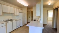 This two-floor residential space offers a practical and well-organized layout designed for comfortable everyday living. The kitchen serves as a central feature of the main level, showcasing white cabinetry with light wood trim that provides generous storage space. A peninsula counter extends into the room, offering additional prep space and a natural dividing point between the kitchen and adjacent living areas. White appliances, including a refrigerator, dishwasher, and a coil-top range, are neatly integrated into the layout for a clean, cohesive look. The flooring in the kitchen features neutral tile, which transitions to carpeting in the surrounding areas, adding warmth to the space. Overhead, a flush-mounted fluorescent light fixture provides bright, even illumination throughout the kitchen. A hallway visible from the kitchen leads further into the home, hinting at additional rooms and living spaces spread across both floors. The overall color palette is light and neutral, with soft beige and white tones throughout, contributing to an airy and open atmosphere. The straightforward design prioritizes functionality and ease of use, making this space well-suited for a variety of living arrangements.