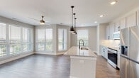 Located at 700 Corporate Center Dr in Raleigh, NC, this thoughtfully designed single-floor residence combines modern finishes with a functional open layout. The heart of the home is a sleek open-concept kitchen and living area, where a granite-topped island with an undermount sink and matte black pendant lighting serves as the central focal point. The kitchen is outfitted with gray shaker-style cabinetry featuring black hardware, granite countertops, a herringbone tile backsplash, and a full suite of stainless steel appliances including a range, microwave, and refrigerator. Wood-look flooring flows consistently throughout the space, lending warmth and continuity. The living area is bathed in natural light through an expansive bank of windows offering outdoor views, complemented by a ceiling fan overhead. The bathrooms are finished with gray vanity cabinetry, granite countertops, large framed mirrors, and black hardware accents. One bathroom includes a soaking tub with floor-to-ceiling marble-style tile surround, while a second features a dedicated vanity area. A spacious walk-in closet provides tiered hanging rods and open shelving for ample storage. The in-unit laundry area accommodates a full-size washer and dryer. A built-in wall shelf in a secondary room adds a practical, custom touch throughout this well-appointed Raleigh residence.