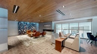 Located at 850 Pacific St in Stamford, Connecticut, this single-floor lounge and collaborative workspace offers a sophisticated blend of comfort and modern design. The interior is immediately distinguished by its warm natural wood-planked ceiling, which spans the entire space and lends a welcoming, organic character to the environment.

The main lounge area is anchored by a bold, floor-to-ceiling bookshelf installation filled with colorful books, serving as a dramatic visual focal point. Adjacent to it, a sleek linear fireplace set into a concrete-textured wall adds warmth and ambiance. The seating arrangement here includes tan leather lounge chairs atop a plush shag rug, creating an inviting reading and conversation nook.

A secondary seating zone features contemporary white upholstered chairs arranged around a solid wood communal table, ideal for informal meetings or collaborative work. Floor-to-ceiling glass walls on one side flood this area with natural light while offering views into adjacent spaces, including what appears to be a fitness area and a room with a living green wall.

A dedicated workstation with a sleek white desk, monitor, and leather accent chair provides a more focused work setting near the glass-paneled entry. Warm-toned tile flooring throughout ties the space together, complementing the wood ceiling and neutral-toned furnishings for a cohesive, refined aesthetic.