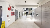 Located on Matteson Avenue in the Mar Vista neighborhood of Los Angeles, this contemporary building's common area presents a thoughtfully designed, open-air lobby and courtyard that sets a clean, modern tone. The ground-floor corridor is finished with large-format light stone tiles underfoot and crisp white walls overhead, creating a bright, welcoming passage that connects the street entrance to the interior courtyard. Exposed concrete beams add an industrial touch, while recessed and track lighting ensure the space remains well-lit throughout.

At the heart of the lobby, a raised white planter hosts a mature tree that rises toward an upper-level balcony and open sky, infusing the space with natural greenery and life. Residents are greeted by a bank of secure package lockers and traditional mailboxes near the entrance, alongside a fire safety panel and utility infrastructure that speak to the building's modern systems. A small bistro table and chairs offer a casual seating nook along the corridor wall.

Beyond the lobby, an expansive tiled terrace opens to the outdoors, bordered by textured block walls with lush plantings and mature trees providing natural privacy. The terrace features contemporary lounge seating, large decorative planters, and steel railings, all bathed in abundant Southern California sunlight — making it a welcoming outdoor retreat for building residents.