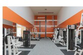 This fitness center in Riverdale, Georgia offers a comprehensive workout facility spread across a single floor, thoughtfully divided into distinct training zones. The space is immediately striking thanks to its bold orange accent walls adorned with motivational phrases such as "ENERGY," "FIT," "GRIT," "#SWEAT," and "DETERMINATION," creating an energizing atmosphere designed to inspire peak performance.

The strength training area is generously sized and outfitted with a wide array of selectorized cable machines, free-weight stations, Smith machines, squat racks, and benches positioned on protective rubber floor mats. Floor-to-ceiling mirrors line the walls throughout this zone, enhancing the sense of openness while allowing for proper form monitoring during exercises.

A separate cardio zone provides a calm contrast, featuring warm neutral tones, crown molding, recessed lighting, and large windows that invite natural light into the space. A lineup of treadmills and elliptical machines is arranged along mirrored walls, creating a bright and airy environment ideal for endurance training.

The facility benefits from soaring ceilings in the main training area, contributing to a spacious and open feel. Durable modular carpet tile flooring spans throughout, offering both comfort and practicality. Overall, the fitness center presents a well-organized, fully equipped environment suited for a broad range of fitness goals.