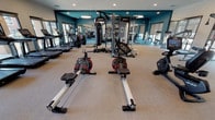 This fitness center offers a comprehensive workout facility designed to accommodate a wide variety of exercise routines. The space is finished with a neutral, textured floor that provides both comfort and durability underfoot, complemented by bold teal accent walls that give the room a modern, energizing aesthetic. Recessed lighting throughout the ceiling ensures the space is well-lit, while large windows along the perimeter flood the interior with natural light and offer views to the outdoors.

The cardio section is prominently featured and includes rowing machines, upright stationary bikes, treadmills, and stair climbers, providing ample options for aerobic training. At the center of the room, a multi-station cable and functional training rig serves as the focal point for strength and resistance work. Additional strength training equipment and free weight stations are visible along the walls, rounding out the facility's offerings.

Full-length mirrors line key wall sections, enhancing the sense of openness and allowing users to monitor their form during workouts. Flat-screen monitors are mounted near the mirrored walls, adding an entertainment or instructional element. The overall layout is open and well-organized, allowing for comfortable movement between equipment stations and accommodating multiple users simultaneously.