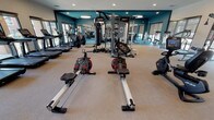 This fitness center offers a comprehensive workout facility designed to accommodate a wide variety of exercise routines. The space is finished with a neutral, textured floor that provides both comfort and durability underfoot, complemented by bold teal accent walls that give the room a modern, energizing aesthetic. Recessed lighting throughout the ceiling ensures the space is well-lit, while large windows along the perimeter flood the interior with natural light and offer views to the outdoors.

The cardio section is prominently featured and includes rowing machines, upright stationary bikes, treadmills, and stair climbers, providing ample options for aerobic training. At the center of the room, a multi-station cable and functional training rig serves as the focal point for strength and resistance work. Additional strength training equipment and free weight stations are visible along the walls, rounding out the facility's offerings.

Full-length mirrors line key wall sections, enhancing the sense of openness and allowing users to monitor their form during workouts. Flat-screen monitors are mounted near the mirrored walls, adding an entertainment or instructional element. The overall layout is open and well-organized, allowing for comfortable movement between equipment stations and accommodating multiple users simultaneously.