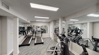 Located at 5445 S Alameda St in Corpus Christi, Texas, this single-floor fitness center is thoughtfully designed to support a full range of workout activities. The space is outfitted with a comprehensive selection of modern exercise equipment, including multiple treadmills with integrated digital displays, elliptical cross-trainers, stationary cycles, and a multi-functional cable strength machine — providing users with both cardiovascular and resistance training options in one convenient facility.

The interior features a neutral, light gray color palette with crisp white trim and columns that lend the space a clean, professional aesthetic. Floor-to-ceiling mirrors are strategically installed along multiple walls, visually expanding the room and allowing users to monitor their form throughout their workouts. The flooring consists of a durable, patterned carpet tile in a blue-gray tone, offering both comfort and practicality underfoot.

Overhead, large flush-mounted fluorescent light panels provide bright, even illumination throughout the space. French-style glass doors allow natural light to filter in and offer views of the exterior surroundings. A water cooler station is conveniently positioned within the facility. The overall ambiance is functional, bright, and well-maintained, making it a practical and welcoming environment for daily fitness use.