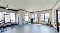 Located at 1971 Willow Loop Way in Knoxville, Tennessee, this single-floor fitness studio offers a well-appointed space designed for a versatile workout experience. The room features clean white walls accented with gray crown molding, and durable gray flooring suited for a variety of exercise activities. Large, multi-pane windows line the walls on multiple sides, flooding the space with natural light and providing views of the surrounding neighborhood. TRX suspension training straps are mounted to the walls, offering users a range of functional strength and resistance training options. Additional equipment includes stability and exercise balls neatly stored on a dedicated rack, as well as a stationary cycle visible along one wall. A ceiling fan is centrally mounted overhead, supporting airflow and comfort during workouts. The open layout allows for ample floor space to accommodate stretching, mat work, or group fitness activities. The overall ambiance is bright, clean, and energizing, making this fitness studio a practical and welcoming environment for residents looking to maintain an active lifestyle.