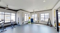 Located at 1971 Willow Loop Way in Knoxville, Tennessee, this single-floor fitness studio offers a well-appointed space designed for a versatile workout experience. The room features clean white walls accented with gray crown molding, and durable gray flooring suited for a variety of exercise activities. Large, multi-pane windows line the walls on multiple sides, flooding the space with natural light and providing views of the surrounding neighborhood. TRX suspension training straps are mounted to the walls, offering users a range of functional strength and resistance training options. Additional equipment includes stability and exercise balls neatly stored on a dedicated rack, as well as a stationary cycle visible along one wall. A ceiling fan is centrally mounted overhead, supporting airflow and comfort during workouts. The open layout allows for ample floor space to accommodate stretching, mat work, or group fitness activities. The overall ambiance is bright, clean, and energizing, making this fitness studio a practical and welcoming environment for residents looking to maintain an active lifestyle.