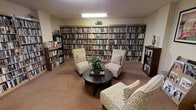 This inviting library is a single-floor space designed for quiet reading and relaxation. The room is dominated by tall, built-in bookshelves that line the walls from floor to near ceiling, housing an extensive and well-organized collection of books spanning multiple genres, including fiction and non-fiction titles. The shelves are densely stocked, offering a rich variety of reading material for visitors.

At the heart of the room, a comfortable seating arrangement encourages leisurely reading and conversation. Two upholstered armchairs with a neutral patterned fabric flank a dark round coffee table, which holds a small potted plant that adds a touch of greenery to the space. A matching sofa is also present, completing the cozy gathering area.

The room features warm carpeting underfoot, contributing to its quiet and comfortable ambiance. Overhead fluorescent lighting provides consistent illumination throughout the space, ensuring the bookshelves and reading area are well-lit. Additional freestanding bookshelves are positioned along the sides of the room, maximizing the available storage for the collection. Overall, this library exudes a warm, welcoming atmosphere well-suited for reading, study, or quiet reflection.