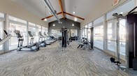 Located at 2890 W 116th Pl in Westminster, Colorado, this well-appointed fitness center offers a comprehensive workout facility designed to meet a wide range of exercise needs. The single-floor space is anchored by soaring vaulted ceilings with exposed wood beams and industrial-style ductwork, giving the interior a modern yet inviting character. Pendant lighting hangs throughout, complementing the abundant natural light that streams in through expansive floor-to-ceiling windows lining multiple walls and offering views of the surrounding outdoor areas.

The main workout floor features herringbone-patterned carpet tile and is equipped with a full lineup of cardio machines, including treadmills and ellipticals, along with cable strength machines, free weights, and resistance equipment. A large mirrored wall anchors one end of the space, enhancing the sense of openness. An adjacent dedicated cycling and functional training studio sets itself apart with a bold, colorful chevron-patterned floor in warm orange, red, and gray tones, a vivid orange accent wall, and a mounted display screen. This area houses stationary bikes, a rowing machine, stability balls, and additional training accessories, creating an energetic and motivating environment for group or individual exercise sessions.