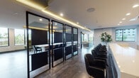 This thoughtfully designed single-floor space blends collaborative and private work environments within a contemporary, open-concept layout. The interior is anchored by sleek glass-partitioned meeting rooms framed in matte black steel, offering visual transparency while maintaining acoustic separation. Dark leather seating lines the corridor leading into these rooms, creating a polished, professional atmosphere complemented by warm cove lighting embedded in recessed ceiling details.

The lounge area is a standout feature, anchored by a generously sized sectional sofa in warm grey, accented with mustard and patterned cushions beneath a cluster of pendant lights with gold-toned interiors. Floor-to-ceiling windows on multiple sides flood the space with natural light and frame views of the surrounding urban landscape and tropical greenery below.

A dedicated quiet zone offers high-backed green velvet pod chairs positioned beside expansive windows overlooking palm-lined outdoor balconies—ideal for focused individual work. The kitchen area features marble-effect countertops, warm wood cabinetry, a professional-grade sink, and wall-mounted screens, combining functionality with refined aesthetics.

Large-format tile flooring in a dark stone finish runs throughout, tying each zone together cohesively. An outdoor terrace with patterned cobblestone paving is accessible via wide sliding glass doors, extending the usable space outdoors.