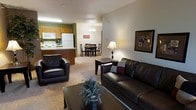 This single-floor space features an open-concept design that integrates the living room, dining area, and kitchen into one cohesive and functional layout. The living room is furnished with dark leather seating, including a sofa and armchair, arranged around a coffee table atop a carpeted floor. Neutral-toned walls and warm lamplight contribute to a relaxed, welcoming ambiance. A framed artwork piece on the wall adds a decorative accent to the space.

The dining area sits just beyond the living room, accommodating a dark wood table with matching chairs beneath a modest chandelier, providing a defined yet open space for meals. The kitchen is positioned adjacent to the dining area and features warm wood cabinetry in a honey-oak finish, white appliances including a refrigerator, microwave, and range, and a raised breakfast bar that visually separates it from the rest of the space. Green accent walls in the kitchen add a subtle pop of color. The overall layout promotes an easy flow between all areas, making the space practical and comfortable for daily living.