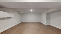 Located at 250 Greene St in New Haven, CT, this loft-to-one-bedroom, one-bathroom residence offers a thoughtfully designed living space with a clean, contemporary aesthetic. Wide-plank hardwood flooring in a warm natural oak tone flows continuously throughout, anchoring the minimalist interior with a sense of warmth and cohesion. The open layout is defined by crisp white walls, generous baseboards, and recessed lighting that keeps the space bright and inviting. The kitchen features a striking peninsula with a wood-tone countertop that mirrors the flooring, complemented by sleek white cabinetry and stainless steel appliances including a built-in oven and range. Structural soffits add architectural dimension to the ceiling plane, creating a layered, loft-inspired character. A dedicated living area benefits from a large black-framed window that draws in natural light and offers views of the surrounding neighborhood's greenery. A wall-mounted smart thermostat and modern control panel reflect the unit's updated, tech-forward conveniences. The overall ambiance is calm and understated, well-suited for those who appreciate a modern, move-in ready home in the heart of New Haven.