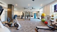 Located at 11005 Octave Dr in Zionsville, Indiana, this fitness center offers a thoughtfully designed, single-floor layout that maximizes functionality and comfort for its users. The main exercise room is generously sized, featuring patterned carpet flooring and a clean, modern aesthetic with recessed lighting and dual ceiling fans that promote air circulation throughout the space. Large floor-to-ceiling windows along one wall flood the room with natural light and offer views of the surrounding greenery, creating an inviting atmosphere for workouts. The exercise area is outfitted with a comprehensive selection of Matrix-brand cardio equipment, including treadmills, ellipticals, stationary bikes, and a multi-station cable machine, complemented by a full rack of free weights and stability balls. Wall-mounted mirrors enhance the sense of openness and allow users to monitor their form. A dedicated wellness room provides a tranquil retreat, appointed with a massage table, an elegantly tiled accent wall in rich blue tones, and a built-in vanity with sink — ideal for recovery and personal care. A carpeted entry corridor connects the main spaces and leads to the restroom, ensuring a smooth and organized flow throughout the facility. The overall design balances practicality with a polished, upscale aesthetic.