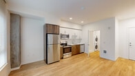 Located at 1670 SW Alder St in Portland's vibrant West End neighborhood, this efficiently designed unit offers a thoughtful, modern layout across a single floor. Light hardwood flooring runs throughout, lending warmth and visual continuity to the open-plan living space. Large windows allow natural light to fill the living area, where an exposed concrete structural column adds an industrial architectural accent alongside clean white walls.

The kitchen is a standout feature, outfitted with a full suite of stainless steel appliances — including a refrigerator, electric range, over-the-range microwave, and dishwasher — paired with two-tone cabinetry combining crisp white upper cabinets and rich wood-toned lower cabinets. White countertops and recessed lighting complete the polished, contemporary look.

Tucked conveniently off the main living area is a stacked washer and dryer unit, offering the practicality of in-unit laundry. The bathroom features a clean, all-white walk-in shower enclosure with chrome fixtures, reflecting the unit's consistent modern aesthetic. Recessed lighting throughout the space ensures a bright and welcoming atmosphere, while the efficient floor plan makes excellent use of every square foot.