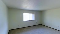 This single-floor space offers a clean, straightforward layout designed for comfortable everyday living. The interior is defined by soft, neutral-toned carpet flooring that extends throughout the main room, providing a warm and quiet underfoot feel. The walls are painted in a light, understated hue that contributes to an open and airy atmosphere. A prominent window serves as a key architectural feature, allowing an abundance of natural light to flood the room and creating a bright, welcoming environment throughout the day. The window is equipped with a horizontal blind, offering practical light control and privacy as needed. The ceiling features a classic textured finish, adding subtle depth and character to the overhead space. Dark baseboard trim runs along the perimeter of the room, providing a clean visual contrast against the lighter walls and carpet. Electrical outlets are thoughtfully positioned along the walls for convenient access. The overall ambiance of the space is calm and neutral, offering a blank canvas well-suited for a variety of uses and personal styling preferences. The single-floor layout ensures easy accessibility and a cohesive flow throughout the space.