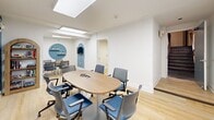 Located at 1228 Spruce Street in Philadelphia's vibrant Center City neighborhood, this single-floor office suite offers a well-designed, functional workspace across two distinct areas. The main conference room serves as the centerpiece of the suite, featuring a large oval wood-topped table surrounded by blue and gray ergonomic rolling chairs, ideal for collaborative meetings or team gatherings. A full-size whiteboard mounted on a striking blue accent wall provides a practical focal point for presentations and brainstorming sessions. The room is brightened by recessed flat-panel ceiling lights, while light wood flooring adds warmth throughout. A decorative arched niche with shelving and a circular accent wall detail lend character and visual interest to the space. Adjacent to the conference room is a compact private office, thoughtfully appointed with a rectangular wood desk, two ergonomic chairs, wall-mounted whiteboard, floating shelves for books, and a blue diamond accent on the wall. Both spaces share a cohesive design palette of white walls, blue accents, and natural wood tones, creating a clean, professional, and inviting atmosphere suited for focused work and productive collaboration.