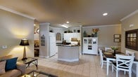 Located at 100 Delaney Dr in Jacksonville, NC, this single-story home offers a well-designed, open-concept layout that seamlessly connects the kitchen, dining, and living areas. The kitchen is a standout feature, showcasing white cabinetry that extends to the ceiling, dark countertops, and a central island that provides additional workspace and casual seating. White appliances, including a full-size refrigerator and microwave, complement the clean, classic aesthetic. Decorative columns frame the kitchen space, adding architectural character and a sense of definition between rooms. Crown molding throughout enhances the refined finish of the interiors. Recessed lighting and overhead fixtures provide consistent, warm illumination across the open floor plan. Light-toned tile flooring runs throughout, contributing to a bright and cohesive feel. A dedicated dining area with a wooden table and white chairs sits adjacent to the kitchen, and a white hutch with wine storage adds both function and charm. A glimpse into a cozy sitting area with natural light from a circular window rounds out this thoughtfully arranged home.
