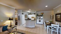 Located at 100 Delaney Dr in Jacksonville, NC, this single-story home offers a well-designed, open-concept layout that seamlessly connects the kitchen, dining, and living areas. The kitchen is a standout feature, showcasing white cabinetry that extends to the ceiling, dark countertops, and a central island that provides additional workspace and casual seating. White appliances, including a full-size refrigerator and microwave, complement the clean, classic aesthetic. Decorative columns frame the kitchen space, adding architectural character and a sense of definition between rooms. Crown molding throughout enhances the refined finish of the interiors. Recessed lighting and overhead fixtures provide consistent, warm illumination across the open floor plan. Light-toned tile flooring runs throughout, contributing to a bright and cohesive feel. A dedicated dining area with a wooden table and white chairs sits adjacent to the kitchen, and a white hutch with wine storage adds both function and charm. A glimpse into a cozy sitting area with natural light from a circular window rounds out this thoughtfully arranged home.