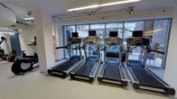 This fitness facility offers a clean, modern environment designed to support a focused and effective workout experience. The space is outfitted with a row of commercial-grade treadmills, each equipped with individual digital display consoles, positioned along a wall of large floor-to-ceiling windows. These expansive windows flood the interior with natural light and provide open views of the surrounding outdoor environment, creating an energizing atmosphere for exercise. The flooring throughout is a durable, cushioned surface well-suited for a high-traffic fitness setting. Overhead, fluorescent lighting complements the abundant natural light, ensuring the space remains bright and well-illuminated at all times. The interior walls are finished in a clean white palette, contributing to the bright and airy feel of the room. The layout is thoughtfully organized to allow ample space between machines, promoting comfort and ease of movement. The single-floor facility presents a straightforward and accessible design, making it a practical and welcoming space for daily fitness activity.