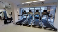 This fitness facility offers a clean, modern environment designed to support a focused and effective workout experience. The space is outfitted with a row of commercial-grade treadmills, each equipped with individual digital display consoles, positioned along a wall of large floor-to-ceiling windows. These expansive windows flood the interior with natural light and provide open views of the surrounding outdoor environment, creating an energizing atmosphere for exercise. The flooring throughout is a durable, cushioned surface well-suited for a high-traffic fitness setting. Overhead, fluorescent lighting complements the abundant natural light, ensuring the space remains bright and well-illuminated at all times. The interior walls are finished in a clean white palette, contributing to the bright and airy feel of the room. The layout is thoughtfully organized to allow ample space between machines, promoting comfort and ease of movement. The single-floor facility presents a straightforward and accessible design, making it a practical and welcoming space for daily fitness activity.