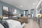 This contemporary single-floor residence offers a thoughtfully designed layout that seamlessly integrates living, dining, and kitchen spaces into a bright, open-concept floor plan. The living area is a standout, featuring dramatic floor-to-ceiling windows with black framing that draw in abundant natural light and offer views of the surrounding architecture. A bold teal accent wall anchors the room, complementing the light hardwood flooring that runs throughout the space. The kitchen is well-appointed with grey shaker-style cabinetry, a herringbone tile backsplash, a suite of stainless steel appliances including a gas range, built-in microwave, dishwasher, and refrigerator, as well as a deep undermount sink. A compact dining nook adjacent to the kitchen features a live-edge wood table illuminated by warm lighting and vibrant artwork, adding a creative touch to the space. The bathroom is clean and spacious, finished in crisp white with a wide vanity, a glass-enclosed tiled shower, and a gallery wall of black-and-white framed photographs that lend a sophisticated, curated feel. Light wood flooring and recessed lighting carry through the bathroom, maintaining design cohesion. Lush greenery, including snake plants, is integrated throughout to enhance the welcoming, modern ambiance.