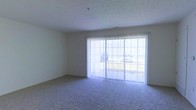 This single-floor space features a carpeted living area that serves as its central focus. The room is finished with neutral-toned walls and a textured ceiling, contributing to a clean and understated aesthetic. A large sliding glass door fitted with vertical blinds dominates one wall, allowing an abundance of natural light to fill the space while providing direct access to an outdoor area. The carpet throughout is a mid-tone gray, offering a practical and comfortable underfoot surface. The overall layout is open and unfurnished, presenting a blank canvas well-suited for a variety of furniture arrangements. The simplicity of the design and the neutral color palette create a versatile environment that can be easily personalized.