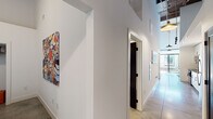 Located at 701 Easley Bridge Road in Greenville, South Carolina, this single-floor unit showcases a distinctive industrial-modern aesthetic defined by dramatic ceiling heights, exposed structural columns finished in dark metal, and polished concrete flooring that flows throughout. The open living area makes a strong architectural statement with its towering white walls, which provide an ideal backdrop for art display, and large windows that draw in natural light. A bold structural column anchors the room, complementing the industrial character of the space.

The kitchen is outfitted with white cabinetry extending floor to ceiling, dark quartz countertops, stainless steel appliances, a deep undermount sink with a matte black faucet, and a striking penny-tile backsplash that adds textural depth. The bedrooms continue the soaring ceiling heights, creating an airy, loft-like atmosphere, with polished concrete and area rugs underfoot.

Two bathrooms feature white floating vanities, dark quartz countertops, matte black hardware, large mirrors with vanity lighting, and tile flooring. The primary bathroom includes a walk-in glass shower with white subway tile surround. A well-lit corridor connects the rooms, with clean white walls that enhance the sense of openness throughout.