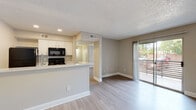 This single-floor unit offers a well-organized, open-concept layout that combines the kitchen and living area into one cohesive space. The kitchen is equipped with white shaker-style cabinetry, light speckled countertops, a peninsula with a stainless steel sink, and a full suite of black appliances including a refrigerator, range, and over-the-range microwave. Light wood-tone flooring runs throughout the main living areas, complementing the neutral warm-beige walls and white baseboards. A textured popcorn ceiling and recessed lighting add depth overhead. The living area is bright and open, anchored by a sliding glass door that leads to a private outdoor balcony with views of the surrounding landscape, allowing ample natural light to fill the space. The bedroom is generously sized, featuring a large window with blinds for natural light and privacy, a ceiling fan, and direct access to a closet. The bathroom includes a full tub and shower combination with white tile surround, wood-tone flooring, and clean, minimal finishes. The entry area provides a welcoming transition into the unit with light flooring and neutral tones carried consistently throughout. Overall, the space presents a clean, functional, and move-in ready interior.