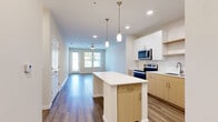 This single-floor residence is designed with a clean, contemporary aesthetic that prioritizes open, functional living. The kitchen serves as a central hub, featuring a two-tone cabinetry scheme that pairs light wood lower cabinets with crisp white upper cabinets and open shelving. A central island with a white countertop provides additional workspace and casual seating, while pendant lighting with brass accents adds a refined touch overhead. Stainless steel appliances, including a refrigerator, range, microwave, and dishwasher, complete the well-equipped space.

The kitchen flows seamlessly into a generously sized living area bathed in natural light from large windows and a glass-paneled door. A ceiling fan serves the living space, and recessed lighting is present throughout. Hardwood-style flooring in warm, neutral tones runs consistently across all rooms, lending cohesion to the layout.

The bedroom is a calm, well-proportioned space with its own ceiling fan and access to a walk-in closet outfitted with wire shelving systems. The bathroom features a soaking tub with subway tile surround, light wood cabinetry with a white countertop, and matching hardwood-style flooring, creating a consistent and polished finish throughout the residence.