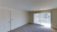 This single-floor space features a straightforward, functional layout centered around a bright and open living area. The room is finished with neutral-toned, wall-to-wall carpeting that provides a clean and understated foundation, complementing the crisp white walls throughout. A sliding glass door serves as a prominent architectural feature, fitted with vertical blinds that allow for adjustable light control. The door frames a view of dense, verdant foliage outside, bringing a natural element into the interior and allowing ample natural light to filter into the room. The ceiling is smooth and painted white, contributing to an overall sense of openness and airiness. The walls are largely unadorned, offering flexibility for personalization and furnishing arrangements. Electrical outlets are positioned at standard intervals along the walls, ensuring practical usability of the space. The overall ambiance is calm and neutral, making it a versatile blank canvas suited to a variety of interior styles and functional uses.