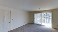 This single-floor space features a straightforward, functional layout centered around a bright and open living area. The room is finished with neutral-toned, wall-to-wall carpeting that provides a clean and understated foundation, complementing the crisp white walls throughout. A sliding glass door serves as a prominent architectural feature, fitted with vertical blinds that allow for adjustable light control. The door frames a view of dense, verdant foliage outside, bringing a natural element into the interior and allowing ample natural light to filter into the room. The ceiling is smooth and painted white, contributing to an overall sense of openness and airiness. The walls are largely unadorned, offering flexibility for personalization and furnishing arrangements. Electrical outlets are positioned at standard intervals along the walls, ensuring practical usability of the space. The overall ambiance is calm and neutral, making it a versatile blank canvas suited to a variety of interior styles and functional uses.