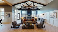 Located in Lombard, Illinois, this single-floor amenity space is a thoughtfully designed clubhouse that blends contemporary style with welcoming functionality. The entry corridor sets an immediate tone of sophistication, featuring herringbone-patterned light wood flooring, sleek black-framed glass partitions on either side, and a dramatic multi-stem chandelier overhead with a warm wood-paneled ceiling inset above.

The main lounge area commands attention with soaring vaulted ceilings accented by exposed dark structural beams and a striking sputnik-style chandelier. A bold vertical black metal lattice partition serves as a dramatic focal point, visually dividing the lounge from a secondary seating zone while adding architectural depth. Curated furnishings include a neutral sectional with burnt-orange accent pillows, textured armchairs, and a glass-topped industrial coffee table arranged around herringbone hardwood floors. Paneled white wainscoting lines the walls alongside curated artwork and sconce lighting.

A casual café-style area transitions seamlessly from the lounge, featuring high-top tables, bar stools, and a wood-planked ceiling detail, ideal for informal gatherings. A sleek fireplace with a dark fluted surround and mounted television anchor one wall, while large transom windows flood the space with natural light and outdoor views. A shuffleboard table adds a recreational touch to this polished, community-centered environment.