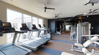 This contemporary fitness center is designed to provide a comprehensive and comfortable workout experience. The space is laid out across a single open floor, maximizing accessibility to a wide range of exercise equipment. Along the perimeter, large floor-to-ceiling windows flood the cardio zone with abundant natural light, creating an energizing atmosphere for users on the treadmills, ellipticals, and other cardio machines arranged in a neat row.

The flooring features a two-tone design with blue and neutral carpet strips, delineating workout zones while adding a sleek, modern aesthetic. Ceiling fans are strategically positioned overhead to ensure consistent airflow throughout the space, contributing to a comfortable training environment alongside recessed lighting.

Toward the rear of the facility, a darker-toned strength and resistance training area is outfitted with a variety of weight machines and equipment, with mirrored walls that visually expand the space and allow exercisers to monitor their form. The contrast between the bright, windowed cardio section and the more focused strength zone creates a functional and dynamic layout. Overall, the fitness center strikes a balance between modern design and practical utility, offering a well-rounded facility suited for diverse fitness routines.