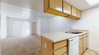 This single-floor residential unit offers a practical and functional layout designed for comfortable everyday living. The open-concept kitchen flows seamlessly into the living area, creating a connected and efficient use of space. The kitchen is outfitted with light wood cabinetry, a countertop peninsula, a stainless steel sink, a dishwasher, a range, and a full-size refrigerator, all set atop tile flooring. Overhead cabinets provide generous storage, while a skylight-style ceiling panel adds brightness to the cooking area. The living room is carpeted and bathed in natural light through a large sliding glass door fitted with vertical blinds, offering access to an outdoor area. White walls and a flush-mount ceiling light give the living space a clean, neutral aesthetic. A dedicated hallway connects the main living areas to a full bathroom and additional storage. The bathroom features tile flooring, a single-sink vanity with wood cabinetry, a wall mirror with overhead lighting, and a full tub-shower combination. A generously sized walk-in closet with wire shelving and carpeted flooring provides ample storage. Throughout the unit, the consistent use of light wood finishes, neutral tones, and tile-and-carpet flooring creates a cohesive and welcoming environment.