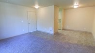 This single-floor residential space offers a straightforward and functional layout designed for comfortable everyday living. The main living area is finished with plush wall-to-wall carpeting, creating a warm and inviting atmosphere, while the entryway and adjoining kitchen zone feature durable tile flooring that transitions seamlessly between spaces. The entry door opens into a modest foyer area that flows naturally into the open living room, which benefits from a clean, uncluttered design with neutral-toned walls throughout.

A partial wall divides the living area from the kitchen corridor, where light-colored cabinetry is visible, suggesting a compact yet practical kitchen setup. The space is equipped with a floor-mounted heating vent, ensuring year-round climate comfort. Overhead lighting fixtures provide consistent illumination across the interior. The neutral color palette of beige and cream tones throughout the walls and flooring lends the space a bright, airy feel, making it versatile for a variety of furnishing styles. Overall, the unit presents a clean, move-in ready environment with a simple and efficient single-floor plan.