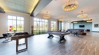 Located at 3175 E Corporate Center Pkwy in Kissimmee, Florida, this sophisticated single-floor clubroom offers a generously sized recreational and social gathering space. The open-concept layout is anchored by wood-look tile flooring laid in a striking herringbone pattern in select areas, complemented by rich hardwood-style planks throughout the main game zone. Soaring ceilings with varied tray and coffered details lend an airy, upscale atmosphere, while a series of elegant chandelier fixtures — including large drum and ornate gold pendant styles — cast warm, inviting light across the space.

Floor-to-ceiling gridded windows line the exterior walls, flooding the interior with natural light and providing expansive views of the surrounding greenery and landscaped grounds. The game area is well-appointed with a full-size billiards table, foosball table, and shuffleboard table, making it a hub for recreation and socializing. A dedicated lounge zone features rich leather armchairs and a textured whitewashed brick accent wall with a wall-mounted television, creating an intimate retreat. A built-in banquette with bold blue upholstered seating adds a design-forward touch near the interior glazed partition wall, while a bar-height dining area rounds out the amenity offerings.