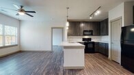 Located at 3701 N Jackson Rd in McAllen, Texas, this single-floor unit offers a well-designed, open-concept layout that seamlessly connects the kitchen and living areas. The kitchen is a standout feature, showcasing rich dark espresso cabinetry with sleek hardware, light quartz-style countertops, a tiled backsplash, and a suite of black appliances including a range, microwave, refrigerator, and dishwasher. A central island with a built-in sink and pendant lighting above adds both functionality and style, while track lighting illuminates the space effectively. Wood-look plank flooring runs throughout the entire unit, providing a warm and cohesive aesthetic. The living area is generously proportioned, featuring a ceiling fan and windows with blinds that allow natural light to filter in, along with access to an outdoor area via a glass door. Bedrooms are well-sized with ceiling fans, natural light, and walk-in closets with built-in shelving. The unit includes two bathrooms: one featuring a tub-shower combination with tiled surround, and a primary bath with a walk-in shower, dark vanity cabinetry, and a framed mirror. Additional storage is provided via louvered-door closets near the kitchen entrance.