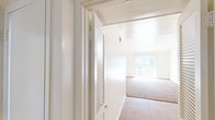 This single-floor residential unit is designed with a clean, neutral aesthetic throughout, creating a bright and airy atmosphere. The living area is generously proportioned, featuring warm beige carpeting, crisp white walls, and a ceiling fan overhead. Natural light flows in through a large window fitted with vertical blinds, illuminating the open layout. A partial-height interior wall defines the transition between the living space and the adjoining areas, adding a subtle architectural detail to the floor plan.

The kitchen is efficiently laid out with wood-look flooring, white cabinetry extending to the ceiling, and a speckled countertop that houses a double stainless steel sink. A coil-top electric range and under-cabinet range hood complete the cooking space, while a window above the counter and a secondary door provide additional natural light and access.

The bedrooms are carpeted and well-lit, each featuring windows with blinds that allow for natural light and privacy. One bedroom benefits from notably tall ceilings, enhancing the sense of space. Louvered closet doors add a classic touch to the storage areas.

The bathroom is finished with wood-look flooring, a long granite-style vanity countertop with a single sink, a wide mirror with overhead lighting, and a shower-tub combination with white tile surround. The overall space is clean, functional, and move-in ready.