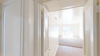 This single-floor residential unit is designed with a clean, neutral aesthetic throughout, creating a bright and airy atmosphere. The living area is generously proportioned, featuring warm beige carpeting, crisp white walls, and a ceiling fan overhead. Natural light flows in through a large window fitted with vertical blinds, illuminating the open layout. A partial-height interior wall defines the transition between the living space and the adjoining areas, adding a subtle architectural detail to the floor plan.

The kitchen is efficiently laid out with wood-look flooring, white cabinetry extending to the ceiling, and a speckled countertop that houses a double stainless steel sink. A coil-top electric range and under-cabinet range hood complete the cooking space, while a window above the counter and a secondary door provide additional natural light and access.

The bedrooms are carpeted and well-lit, each featuring windows with blinds that allow for natural light and privacy. One bedroom benefits from notably tall ceilings, enhancing the sense of space. Louvered closet doors add a classic touch to the storage areas.

The bathroom is finished with wood-look flooring, a long granite-style vanity countertop with a single sink, a wide mirror with overhead lighting, and a shower-tub combination with white tile surround. The overall space is clean, functional, and move-in ready.