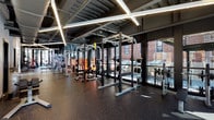 This expansive fitness center is thoughtfully organized into distinct workout zones, each designed to support a comprehensive range of training activities. The space features an industrial aesthetic with exposed steel structural elements, dark painted ceilings, and linear LED lighting running diagonally overhead, creating a dynamic and energized atmosphere throughout.

The strength training area is outfitted with multiple power racks, cable machines, barbells, weight benches, and plate storage, all set on durable rubber flooring. Adjacent to this zone, a dedicated cardio section lines the perimeter wall, featuring treadmills, ellipticals, and rowing machines positioned directly in front of expansive floor-to-ceiling windows. These large glass panels flood the space with natural light and offer views of the surrounding brick urban streetscape.

A separate studio area with hardwood flooring provides open space for functional training and flexibility work, equipped with stability balls and balance tools. Perforated metal partitions divide zones while maintaining visual openness. A stainless steel elevator provides convenient access, and large glass doors lead directly to an outdoor terrace with seating and greenery, seamlessly connecting the interior fitness space to the outdoors.