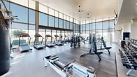 This well-appointed fitness center is designed to deliver a premium workout experience within a bright, open, and architecturally impressive space. Soaring double-height ceilings and expansive floor-to-ceiling glass curtain walls define the facility's most striking architectural feature, flooding the interior with abundant natural light and offering sweeping panoramic views of the surrounding cityscape and landscape.

The cardio zone is generously equipped with multiple treadmills, elliptical trainers, and rowing machines—including WaterPower rowers—all positioned to take full advantage of the view. Ceiling fans overhead ensure comfortable airflow throughout. The strength training area is equally comprehensive, featuring a full rack of dumbbells spanning a wide weight range, adjustable benches, cable machines, and a multi-functional cable crossover station. Rubberized flooring in the free weights area provides safety and durability.

Mirrored walls in the strength section enhance the sense of space and support proper form during workouts. Wall-mounted flat-screen televisions are positioned for easy viewing from multiple areas of the floor. A dedicated service area with cabinetry and a water station adds convenience for users. The overall ambiance is clean, modern, and motivating, combining functional design with an energizing atmosphere suited for a full range of fitness activities.
