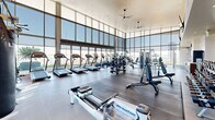 This well-appointed fitness center is designed to deliver a premium workout experience within a bright, open, and architecturally impressive space. Soaring double-height ceilings and expansive floor-to-ceiling glass curtain walls define the facility's most striking architectural feature, flooding the interior with abundant natural light and offering sweeping panoramic views of the surrounding cityscape and landscape.

The cardio zone is generously equipped with multiple treadmills, elliptical trainers, and rowing machines—including WaterPower rowers—all positioned to take full advantage of the view. Ceiling fans overhead ensure comfortable airflow throughout. The strength training area is equally comprehensive, featuring a full rack of dumbbells spanning a wide weight range, adjustable benches, cable machines, and a multi-functional cable crossover station. Rubberized flooring in the free weights area provides safety and durability.

Mirrored walls in the strength section enhance the sense of space and support proper form during workouts. Wall-mounted flat-screen televisions are positioned for easy viewing from multiple areas of the floor. A dedicated service area with cabinetry and a water station adds convenience for users. The overall ambiance is clean, modern, and motivating, combining functional design with an energizing atmosphere suited for a full range of fitness activities.