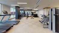 This modern fitness center offers a comprehensive workout environment spread across a single, open floor. The space is thoughtfully designed with a clean, contemporary aesthetic, featuring light neutral tile flooring, crisp white walls accented by dark decorative panels, and a combination of recessed linear and circular pendant lighting that creates a bright, energizing atmosphere.

The cardio zone is positioned along a windowed wall, where Matrix treadmills and other cardio equipment face large floor-to-ceiling windows overlooking lush palm trees, flooding the area with natural light and scenic outdoor views. Floor-to-ceiling mirrors along multiple walls enhance the sense of spaciousness throughout.

The strength training section is well-appointed with a full rack of dumbbells, cable machines, and selectorized equipment, while a dedicated functional training area provides ample open floor space for stretching, yoga mats, stability balls, BOSU trainers, and a large flat-screen display for guided workouts. TRX suspension trainers and a wall-mounted mat round out the functional zone.

A refined refreshment station features warm wood cabinetry, a stainless steel under-counter refrigerator stocked with bottled water, a water purifier, and open shelving neatly stocked with fresh towels — offering a polished, hospitality-grade touch to the overall fitness experience.