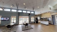 This single-story amenity space is designed to serve as a multifunctional gathering and recreation hub. The layout is open and airy, with two distinct yet connected zones that flow seamlessly across light wood-look flooring.

The social lounge and dining area anchors one side of the space, featuring multiple tables and modern black chairs arranged atop bold black-and-white striped area rugs. A large glass overhead garage door dominates the exterior wall, allowing the space to open fully to the outdoors and flooding the interior with natural light. A row of clerestory windows above further enhances the brightness. A wall-mounted television and a compact kitchenette equipped with a coffee station, light cabinetry, and a dark tile backsplash provide practical amenities for residents and guests.

The recreation area occupies an adjacent, generously proportioned room with soaring vaulted ceilings accentuated by a recessed skylight detail. Clerestory windows wrap the upper walls, bringing in ample daylight. This room includes a ping-pong table, a bar-height work table with stools, a wall-mounted shelf display, a flat-screen television, and a casual seating arrangement with rattan-style chairs. The overall aesthetic throughout is clean and contemporary, combining neutral tones, durable finishes, and thoughtful amenities.