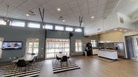 This single-story amenity space is designed to serve as a multifunctional gathering and recreation hub. The layout is open and airy, with two distinct yet connected zones that flow seamlessly across light wood-look flooring.

The social lounge and dining area anchors one side of the space, featuring multiple tables and modern black chairs arranged atop bold black-and-white striped area rugs. A large glass overhead garage door dominates the exterior wall, allowing the space to open fully to the outdoors and flooding the interior with natural light. A row of clerestory windows above further enhances the brightness. A wall-mounted television and a compact kitchenette equipped with a coffee station, light cabinetry, and a dark tile backsplash provide practical amenities for residents and guests.

The recreation area occupies an adjacent, generously proportioned room with soaring vaulted ceilings accentuated by a recessed skylight detail. Clerestory windows wrap the upper walls, bringing in ample daylight. This room includes a ping-pong table, a bar-height work table with stools, a wall-mounted shelf display, a flat-screen television, and a casual seating arrangement with rattan-style chairs. The overall aesthetic throughout is clean and contemporary, combining neutral tones, durable finishes, and thoughtful amenities.
