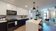 This single-floor residence showcases a thoughtfully designed interior with a cohesive modern aesthetic. The kitchen serves as a central highlight, featuring a striking two-tone cabinet scheme pairing crisp white upper cabinets with deep navy lower cabinets accented by gold hardware. A geometric hexagonal tile backsplash adds visual texture, while white quartz countertops provide a clean, bright contrast. A substantial kitchen island with seating offers casual dining and additional workspace, illuminated overhead by a pair of black industrial-style pendant lights. Stainless and black Whirlpool appliances, including a French door refrigerator, gas range, and built-in microwave, complete the functional kitchen suite. Light wood-tone flooring runs throughout, lending warmth and continuity across the living spaces.

The bedrooms are well-proportioned, each benefiting from floor-to-ceiling windows that invite natural light and provide urban views of the surrounding cityscape. The bathrooms offer distinct personalities: one features a bold emerald green accent wall, large-format tile flooring, and a tub-shower combination, while a second presents a neutral palette with a walk-in shower, wood-toned vanity, and black hardware accents throughout.