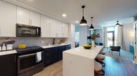 This single-floor residence showcases a thoughtfully designed interior with a cohesive modern aesthetic. The kitchen serves as a central highlight, featuring a striking two-tone cabinet scheme pairing crisp white upper cabinets with deep navy lower cabinets accented by gold hardware. A geometric hexagonal tile backsplash adds visual texture, while white quartz countertops provide a clean, bright contrast. A substantial kitchen island with seating offers casual dining and additional workspace, illuminated overhead by a pair of black industrial-style pendant lights. Stainless and black Whirlpool appliances, including a French door refrigerator, gas range, and built-in microwave, complete the functional kitchen suite. Light wood-tone flooring runs throughout, lending warmth and continuity across the living spaces.

The bedrooms are well-proportioned, each benefiting from floor-to-ceiling windows that invite natural light and provide urban views of the surrounding cityscape. The bathrooms offer distinct personalities: one features a bold emerald green accent wall, large-format tile flooring, and a tub-shower combination, while a second presents a neutral palette with a walk-in shower, wood-toned vanity, and black hardware accents throughout.