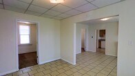 This single-floor space presents a straightforward, functional layout with an open main area that serves as the central hub connecting to surrounding rooms. The flooring throughout the main area consists of light beige ceramic tiles laid in a classic grid pattern, providing a clean and easy-to-maintain surface. The ceiling features a suspended drop-tile grid system with integrated fluorescent lighting panels, offering consistent, even illumination throughout the space.

The walls are finished in a neutral off-white tone, creating a blank canvas adaptable to various uses. Two doorways are visible from the main area — one leading to an adjacent room with darker flooring, and another opening to a secondary space beyond. A partial wall divides portions of the layout, adding subtle definition without fully closing off the flow between areas.

To the right, a glimpse into the kitchen reveals wooden cabinetry in a warm medium-brown tone, light-colored countertops, and what appears to be a dishwasher or white appliance along the base. The kitchen features its own tile flooring that coordinates with the main area. Natural light enters through at least one window visible to the left side of the space, supplementing the overhead lighting and lending a modest sense of openness to the interior.