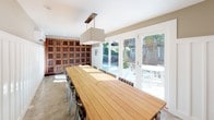 This thoughtfully designed wine room offers a refined space for wine storage and entertaining. The room's most striking feature is the floor-to-ceiling wall of rich wood wine lockers with metal mesh doors, providing secure, organized storage for an extensive collection. A long teak dining table anchors the center of the room, surrounded by woven rattan chairs, creating an inviting setting for tastings and gatherings. A rectangular pendant light fixture hangs above the table, providing focused illumination.

The wet bar area is equipped with white upper cabinets featuring frosted glass doors, dark lower cabinetry, a stainless steel farmhouse sink, and a compact refrigerator—ideal for serving and entertaining. Classic white beadboard wainscoting lines the walls beneath a warm taupe upper wall finish, contributing to the room's clean, polished aesthetic. A wall-mounted mini-split system ensures precise climate control, essential for proper wine storage.

Large glass French doors along one wall flood the space with natural light and open directly to an outdoor patio, seamlessly connecting the interior to the exterior. Polished concrete floors complete the refined yet functional design of this exceptional wine room.